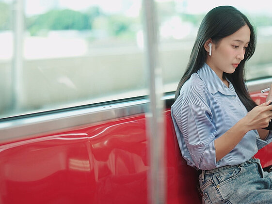A woman sitting on a train with her back to the window. She is wearing earphones while working on her wireless device